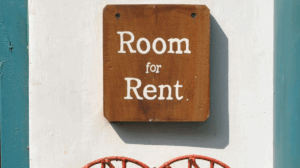 Wooden “Room for Rent” sign on white wall with teal framing and red decoration, symbolising the softening rental market